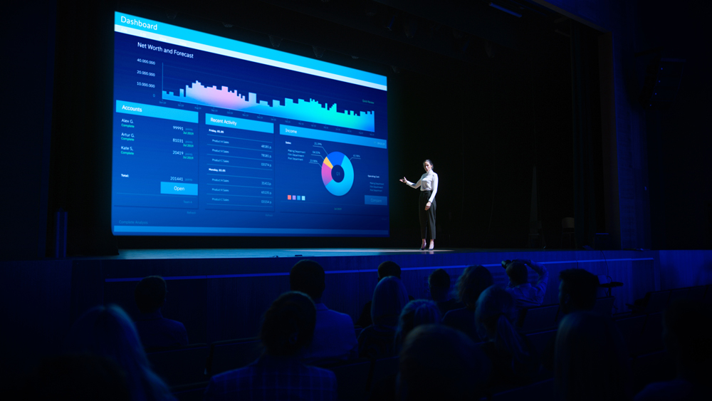 A speaker presenting a powerpoint of a financial dashboard on a large screen to an audience in a darkened lecture hall.
