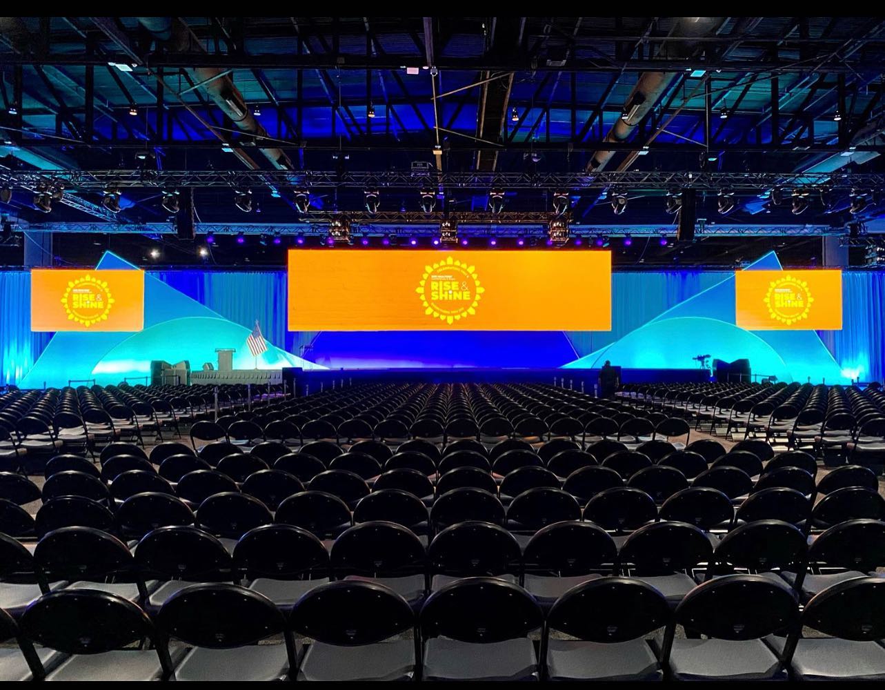 A view of a conference room being prepared, with a vividly colored widescreen at the front and rows of empty chairs arranged for attendees.
