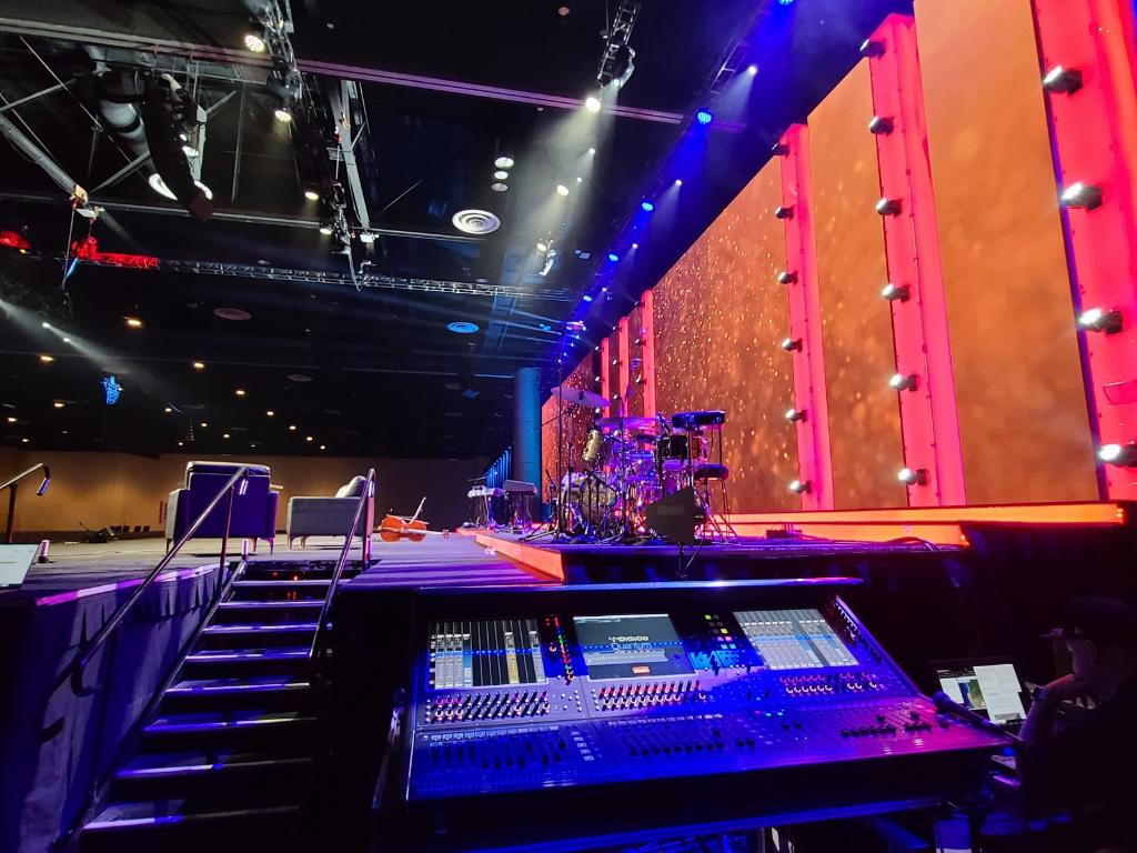 View from behind a sound mixing console at a live event venue, looking towards a brightly lit stage with vibrant red panels and sparkling lights. Musical instruments including a drum set are set up on the stage, with no performers present.