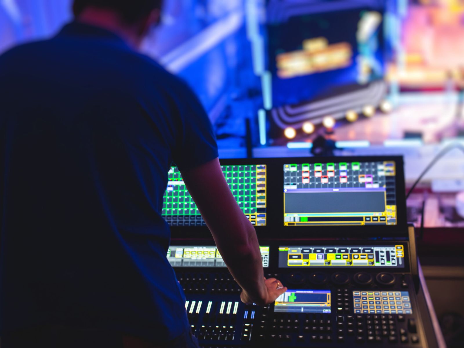 Rear view of an individual using an illuminated sound mixing console in a dark room with screens displaying colorful graphics.