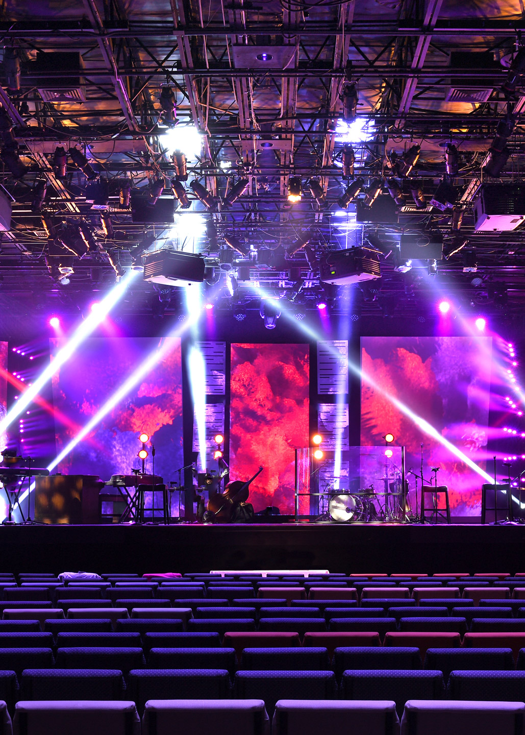 An empty event hall with rows of chairs facing a stage lit with purple lights and screens displaying vibrant, red visuals.