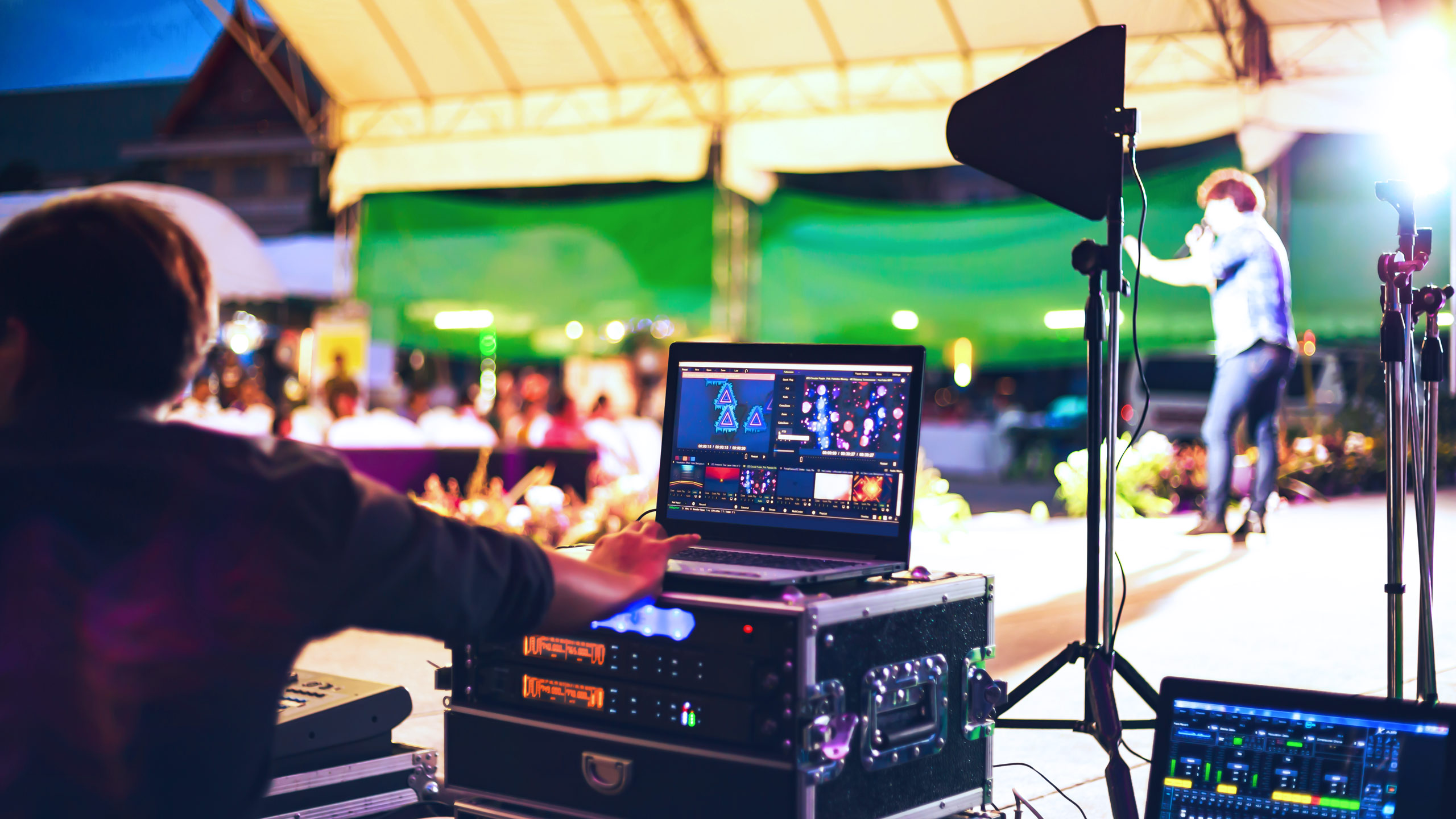 View from behind a technical operator working on a laptop with visual production software, with a blurred background featuring an on-stage performer and an audience at an outdoor event.