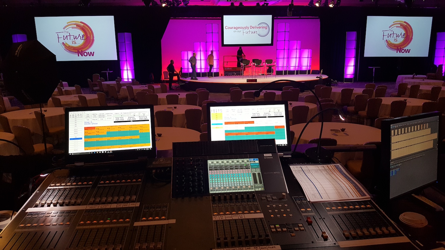 A view from the audio-visual control area of a conference room set up with round tables, a stage, and screens displaying the message 'The Future is... NOW'.