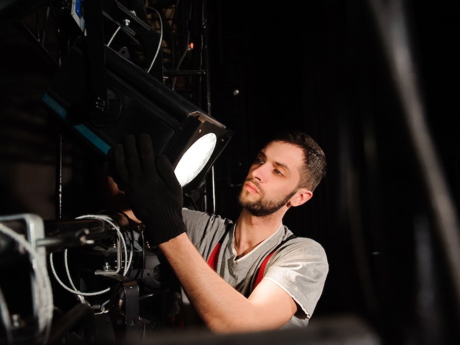 A man adjusting a studio light, wearing gloves and a headset, with rigging equipment in the background.