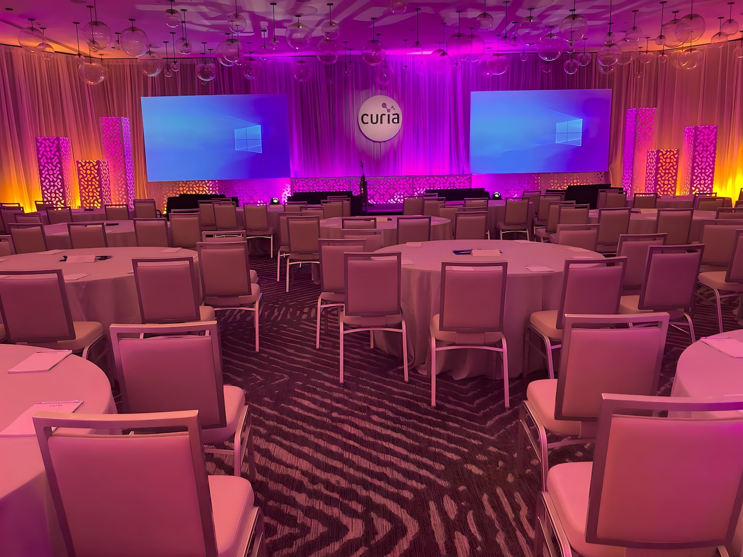 A Miami conference setup with round tables and pink lighting, a stage with a large screen displaying a graphic, and the branding 'curia' prominently featured.
