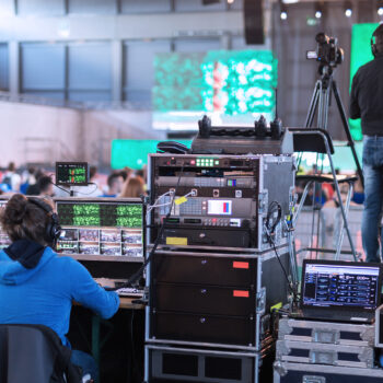 Two people operating broadcasting equipment at a live event, with screens showing the stage and audience, and a camera operator filming nearby.