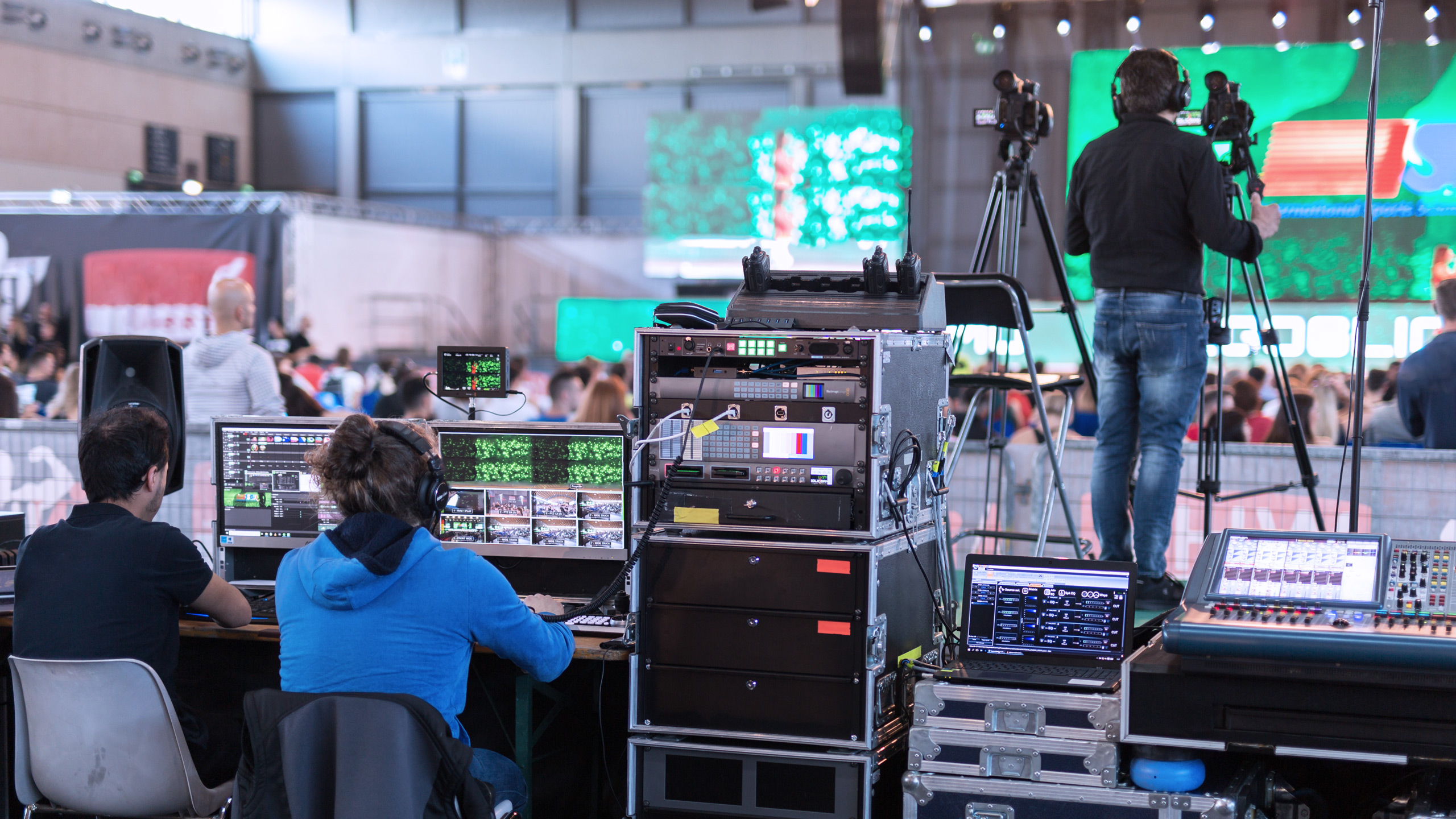 Two people operating broadcasting equipment at a live event, with screens showing the stage and audience, and a camera operator filming nearby.